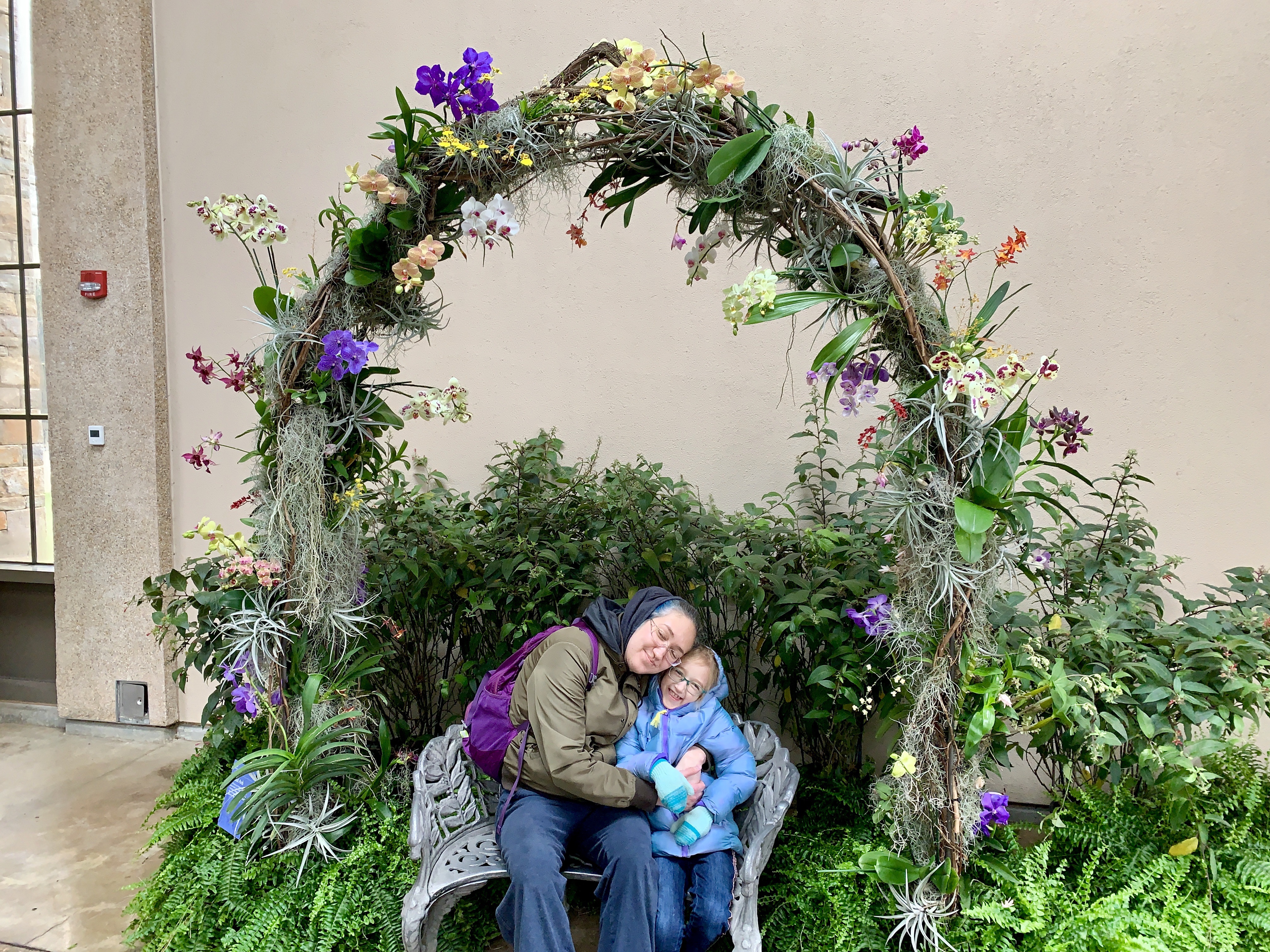 Wife and daughter hugging beneath an arch made of orchids. 